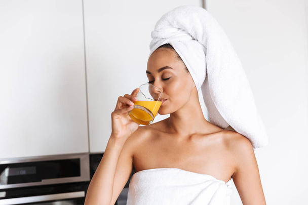 Healthy young asian woman wrapped in a shower towel drinking orange juice from a glass while standing on a kitchen - Φωτογραφία, εικόνα