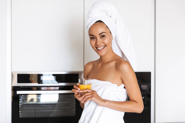 Smiling young asian woman wrapped in a shower towel holding orange juice glass while standing on a kitchen - 写真・画像