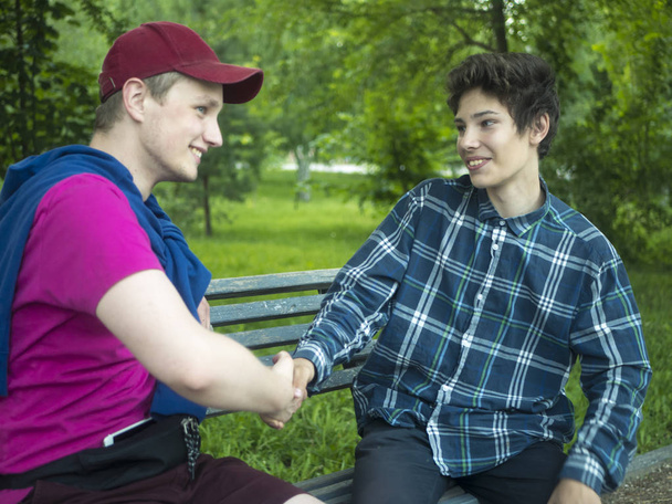 two young smiling friends handshake saluting each other sitting on the bench outdoors on a summer day - Fotoğraf, Görsel