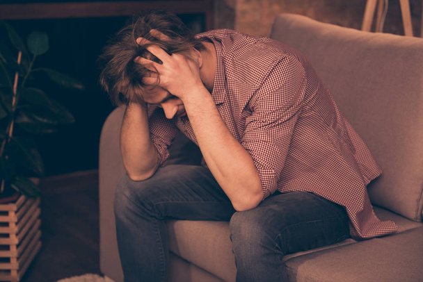 Frustrated young guy with bristle is holding his head, he is upset because his business has failed, sitting on the couch alone, bad luck, relationship problems, suffering from headache - Foto, Imagem