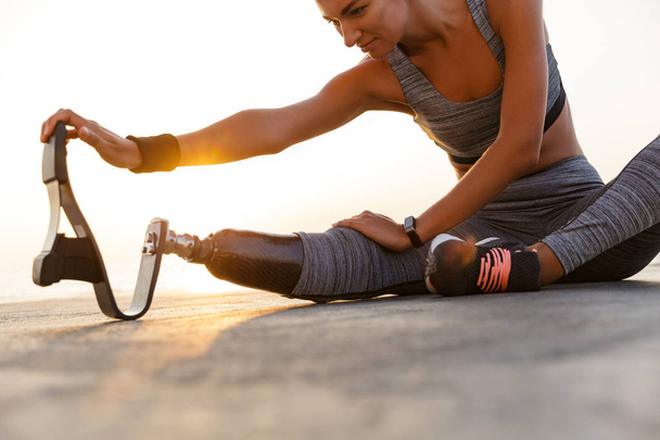 Cropped image of motivated disabled athlete woman with prosthetic leg doing stretching exercises while sitting at the beach - Foto, imagen