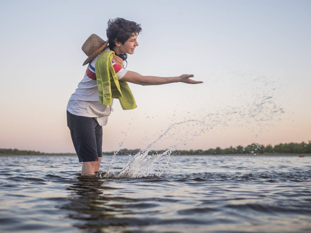 retrato del joven hipster con sombrero y toalla haciendo salpicaduras de agua en agua de mar
 - Foto, Imagen