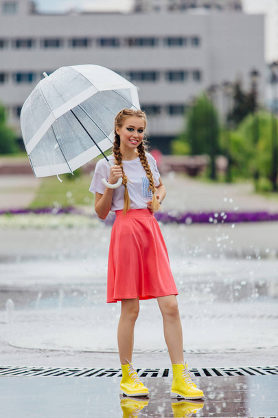 Young pretty girl with two braids in yellow boots and with transparent umbrella stands near fountain. Rainy day in city. - Foto, immagini