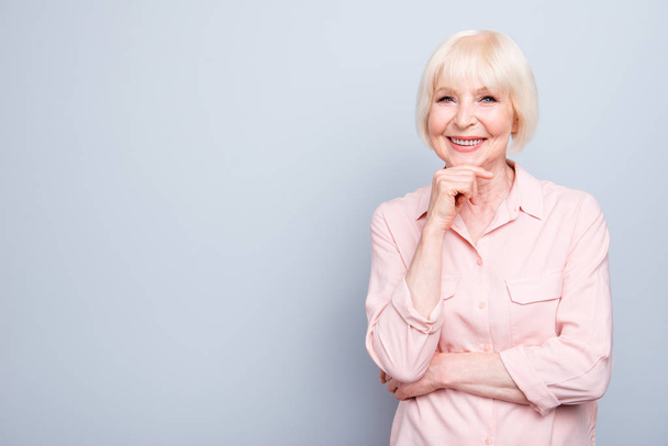 Old blonde lady smiling holding hand under chin on grey background, standing straight, copy space - 写真・画像