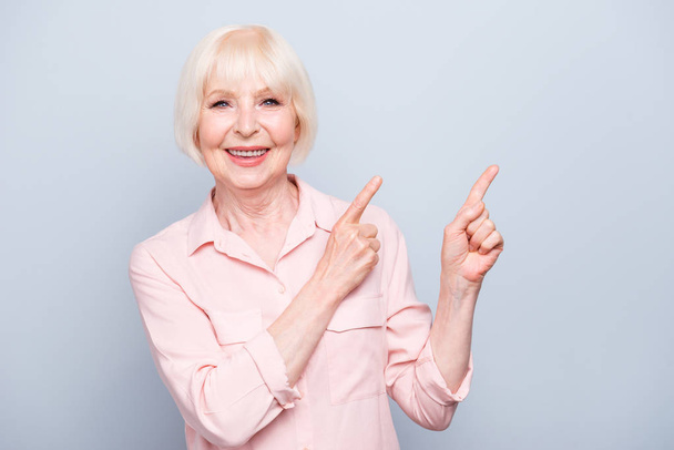 Old blonde glad lady smiling fingers pointing up over grey background, copy space - 写真・画像