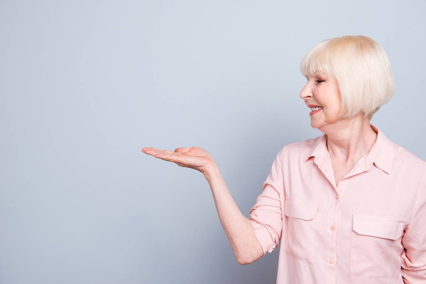 Old adult blonde glad cheerful lady smiling, showing gesture with hand, palm to the right, over grey background, looking right, isolated, copy space - Fotoğraf, Görsel