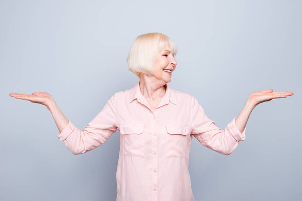Old adult blonde glad cheerful lady smiling, showing gesture with hands, palms two sides, over grey background, looking left, isolated - Foto, Imagen