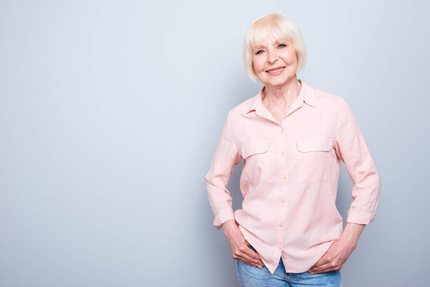 Portrait of old adult blonde glad cheerful caucasian lady smiling, wearing pink shirt and jeans, over grey background, isolated, copy space - Photo, Image