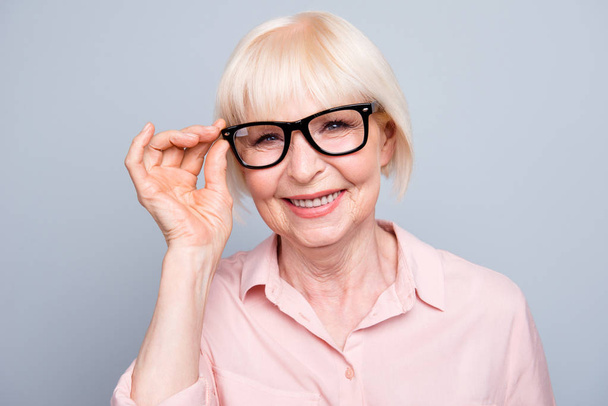 Portrait of old adult blonde glad cheerful caucasian nice lovely lady smiling, wearing eye glasses, showing touch spectacles gesture, over grey background, isolated - Foto, immagini