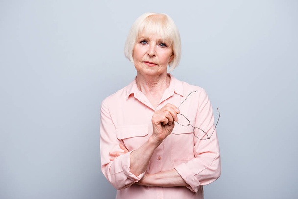 Portrait of old adult blonde caucasian sad serious lady, holding eye glasses, thinking, looking straight over grey background, isolated - Foto, Imagen
