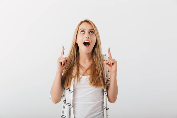 Portrait of a cheerful young casual girl pointing fingers up isolated over white background - Φωτογραφία, εικόνα