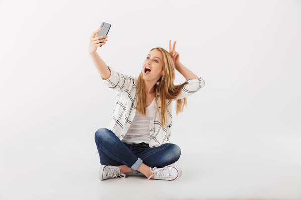 Portrait of cheerful young casual girl taking selfie with mobile phone while sitting with legs crossed isolated over white background - Foto, Imagen