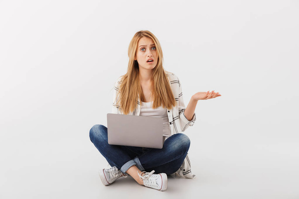 Portrait of confused young casual girl with laptop computer sitting with legs crossed isolated over white background - Fotoğraf, Görsel