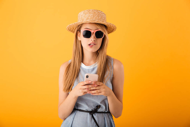 Portrait of a pensive young blonde woman in summer hat and sunglasses using mobile phone isolated over yellow background - Valokuva, kuva