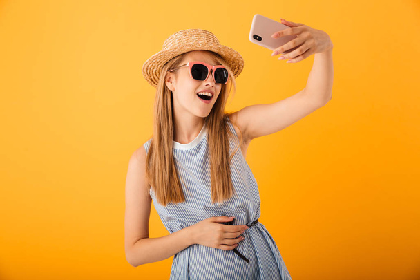 Smiling young blonde woman in summer hat and sunglasses taking a selfie isolated over yellow background - Foto, Bild