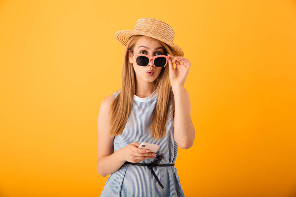 Şaşırmış gibi bir sarışın genç kadın yaz şapka ve güneş gözlüğü izole sarı arka plan üzerinde cep telefonuyla portresi - Fotoğraf, Görsel