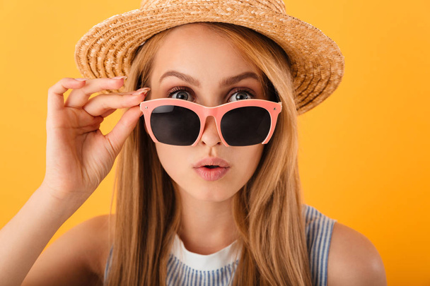 Close up portrait of a pretty young blonde woman in summer hat and sunglasses looking at camera isolated over yellow background - Foto, imagen