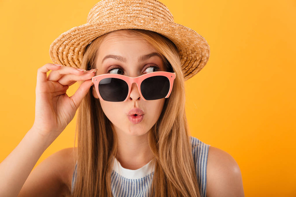 Close up portrait of a pretty young blonde woman in summer hat and sunglasses looking away isolated over yellow background - Foto, Bild