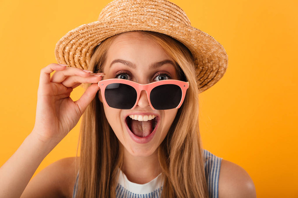Close up portrait of a cheerful young blonde woman in summer hat and sunglasses looking at camera isolated over yellow background - Zdjęcie, obraz
