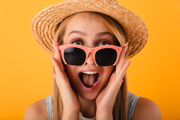 Close up portrait of a happy young blonde woman in summer hat and sunglasses looking at camera isolated over yellow background - Fotografie, Obrázek