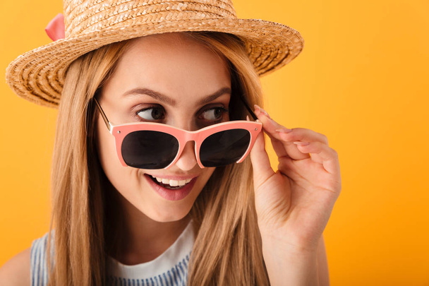Close up portrait of a cheerful young blonde woman in summer hat and sunglasses looking away isolated over yellow background - Fotó, kép