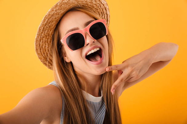 Cheerful young blonde woman in summer hat and sunglasses taking a selfie isolated over yellow background - Foto, imagen