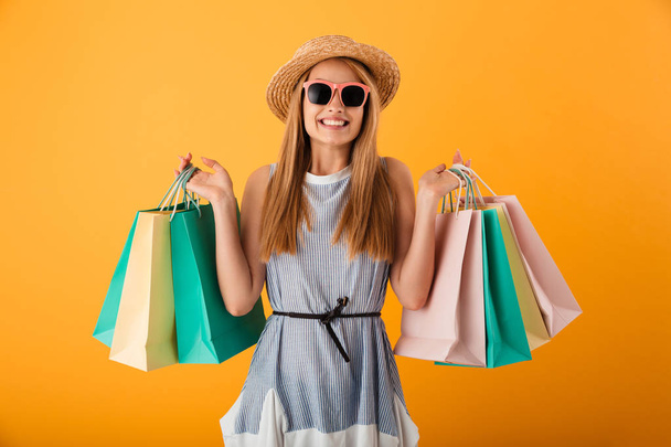 Portrait of a cheerful young blonde woman in summer hat and sunglasses holding shopping bags isolated over yellow background - Фото, зображення