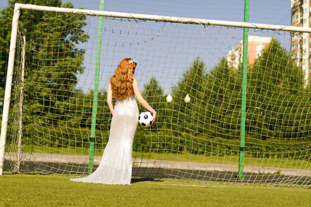 Uma menina com cabelo vermelho fica no gol de futebol de volta com uma bola em um vestido branco
. - Foto, Imagem