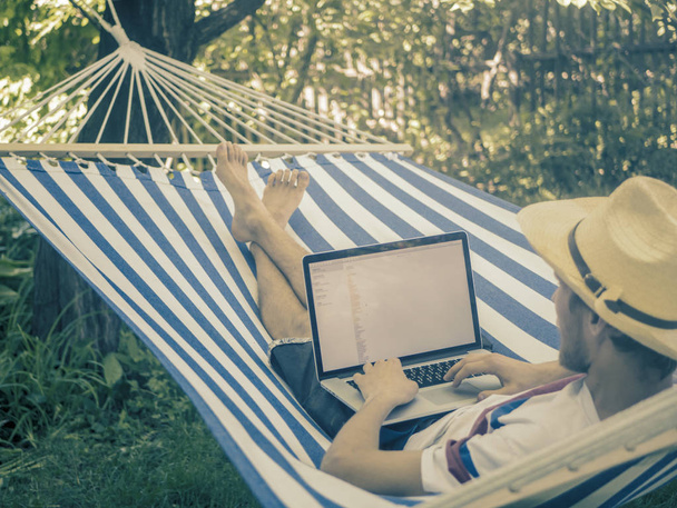 back view of lying young man in hat working with laptop on hammock on a summer day - Foto, Imagen