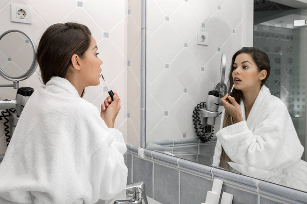 Sivunäkymä tyttö valkoisessa kylpytakissa soveltamalla värikäs huulipuna peilin edessä seisoo valossa kylpytakki
 - Valokuva, kuva