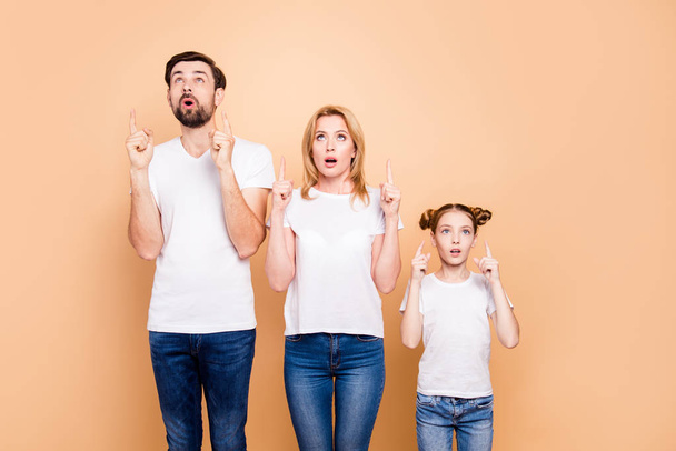 Bearded father, blonde mother and their little daughter standing straight and pointing up with their fingers on beige background mum mommy - Valokuva, kuva