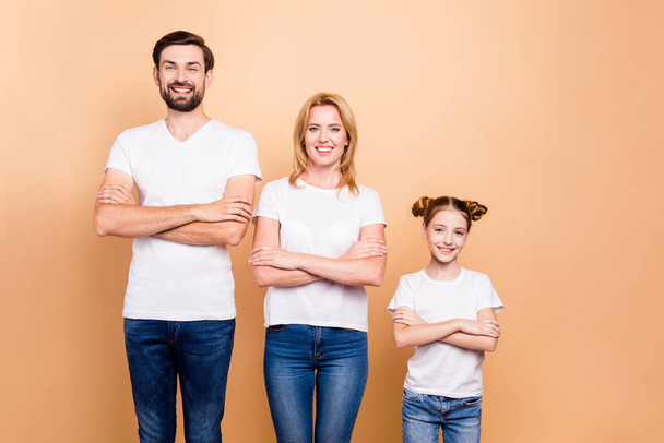 Bearded father, blonde mother and their little daughter wearing blue jeans and white T-shirts standing straight with crossed arms, laughing on beige background - Fotografie, Obrázek
