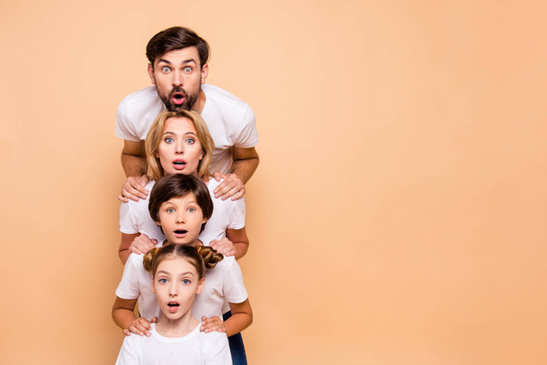 Adorable attractive surprised family, bearded father, blonde mother, boy and girl wearing white T-shirts, standing in odrer of hierarchy, holding hands on each other's shoulders. Copy space - Foto, afbeelding