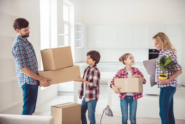 Young happy smiling family four persons unpacking carton packages with stuff in light kitchen, moving to new house - 写真・画像