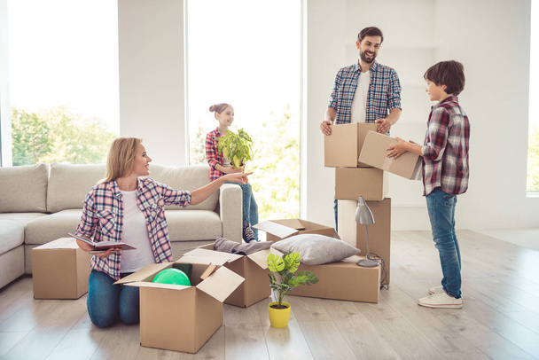 Young happy smiling family four persons unwrapping carton boxes with stuff in light studio living room, moving to new flat, check list - Foto, Bild