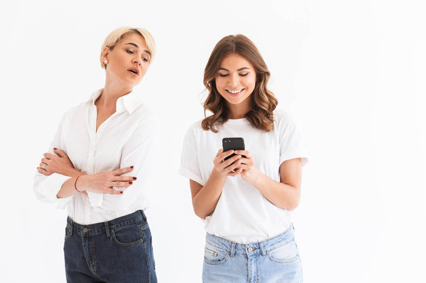 Portrait of mature curious woman peeking at her daughter who holding and using black mobile phone with smile standing isolated over white background - Φωτογραφία, εικόνα