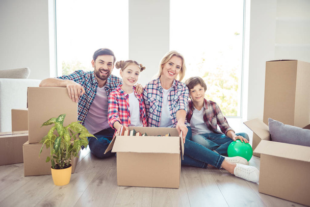 Young happy smiling family four persons sitting on floor unwrapping carton boxes packages with stuff things in light living room, moving to new flat - Foto, Imagem