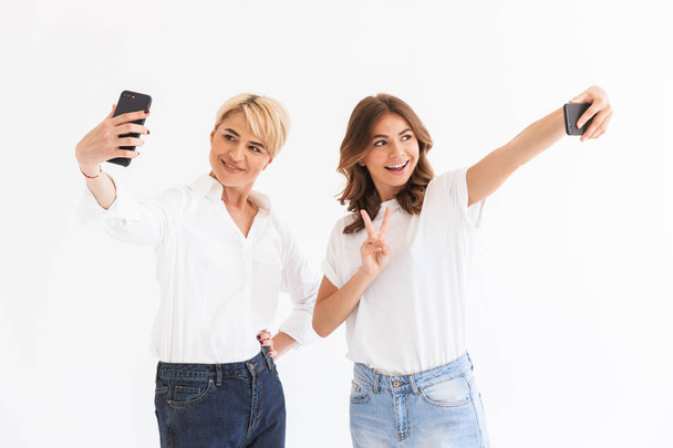 Image of young woman standing with her mother make selfie by mobile phones isolated over white background. - Φωτογραφία, εικόνα