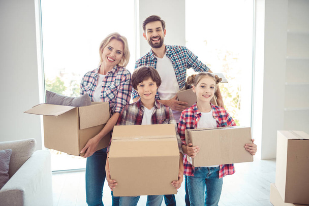 Young happy smiling family four persons wearing casual standing together holding carton boxes with stuff things in light living room moving to new flat - Фото, зображення