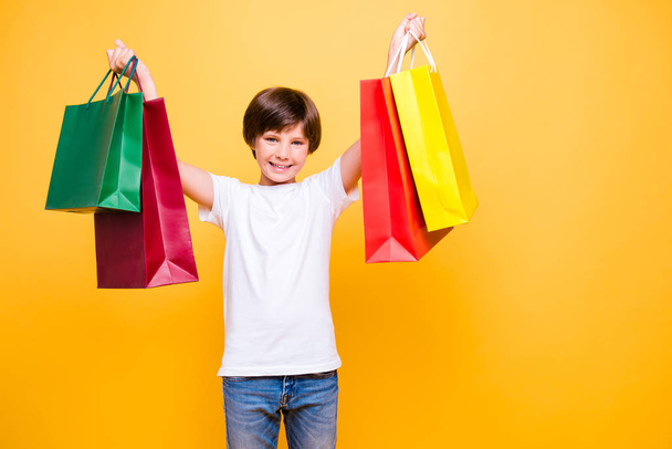 Portrait of attractive young cheerful school boy, smiling standing raising hands up with colorful bags over yellow background, isolated. Copy space - Photo, Image