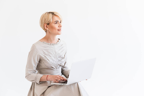 Beautiful middle aged woman with short blond hair wearing dress sitting and using silver laptop isolated over white background in studio - Fotoğraf, Görsel