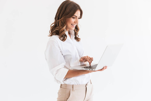 Porträt einer entzückenden lächelnden Frau mit langen braunen Haaren, die einen silbernen Laptop vor weißem Hintergrund im Studio hält - Foto, Bild