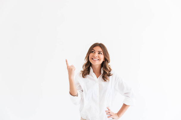 Photo of joyous smiling woman 20s with long brown hair pointing finger upward at copyspace isolated over white background in studio - Photo, Image