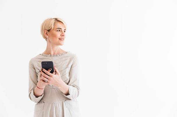 Smiling middle aged woman with short blond hair wearing dress holding black smartphone and looking aside at copyspace isolated over white background in studio - Zdjęcie, obraz