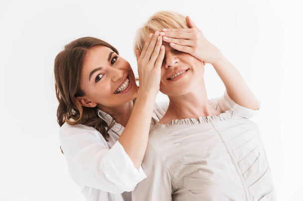 Mooie vrouw 20s bedekking ogen van haar moeder 40s die achter en lachend op camera geïsoleerd op witte achtergrond - Foto, afbeelding