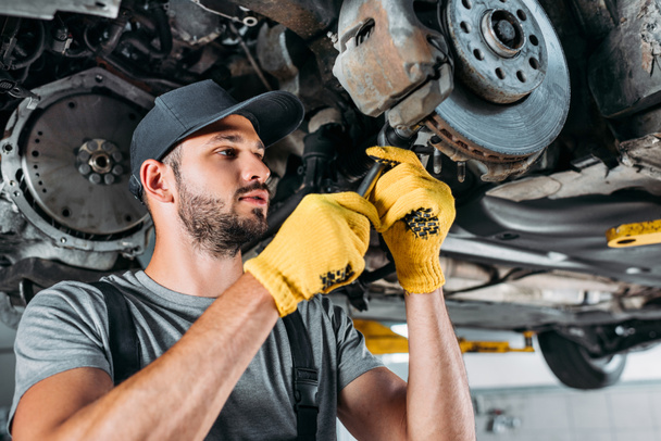 professional mechanic in uniform repairing car without wheel in workshop - Фото, изображение