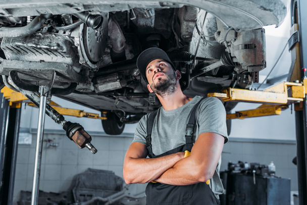 профессиональный рабочий со скрещенными руками, смотрящий на автомобиль в мастерской
 - Фото, изображение