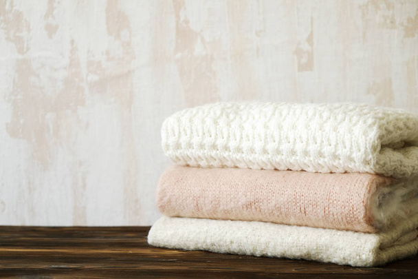 Bunch of knitted warm pastel color sweaters with different knitting patterns folded in stack on brown wooden table, white textured wall background. Fall winter knitwear. Close up, copy space for text. - Фото, изображение