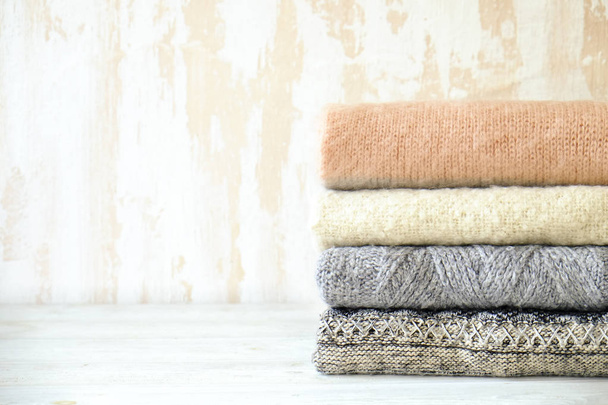 Bunch of knitted warm pastel color sweaters with different knitting patterns folded in stack on white wooden table, textured wall background. Fall winter season knitwear. Close up, copy space for text - Foto, afbeelding