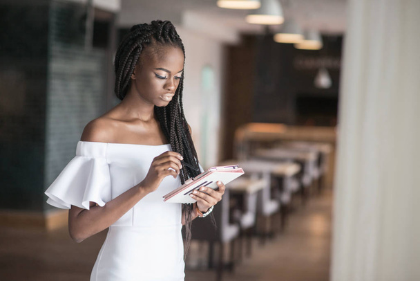 Pretty girl with shiny skin wearing beautiful white dress and standing in a cafe or a restaurant. African american woman with the tablen in her hands. - Fotoğraf, Görsel